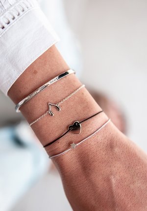 Quatre bracelets sur un poignet : un bracelet ouvert en argent gravé, une chaîne avec un os de vœu en argent, un charme en forme de cœur noir et un charme en forme d'étoile en argent sur une fine chaîne.