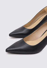 Tacones altos de cuero negro con un diseño de punta afilada, textura suave y un corte bajo en el empeine que deja al descubierto el forro interior color beige.