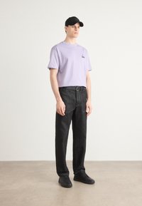Lavendel bomulds t-shirt med lille sort logo, sat sammen med mørkegrå jeans og sorte sneakers. Modellen bærer en sort kasket.