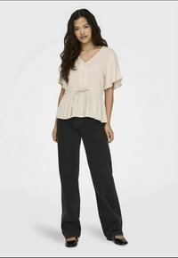 Μπεζ μπλούζα με V-λαιμό, κοντά μανίκια, λεπτομέρεια με κουμπιά και μαζεμένη μέση. Φορεμένη με μαύρα παντελόνια ευρείας γραμμής και μαύρες μπαλαρίνες.