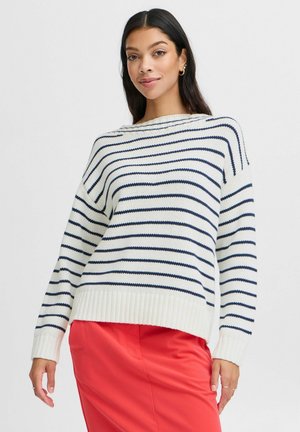 Striped knit sweater in cream with dark blue stripes, featuring a ribbed hem and loose fit. Worn over a red skirt.