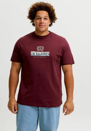 Mand med krøllet hår iført en vinrød Jack & Jones t-shirt og lyseblå denimshorts står foran en ensfarvet baggrund.