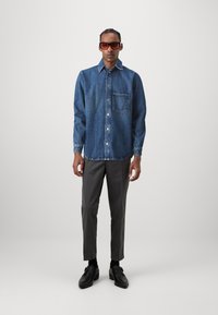 Camicia di denim con chiusura a bottoni, taschino sul petto e colletto a contrasto; pantaloni grigio scuro, scarpe nere e occhiali da sole marroni.