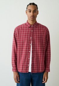 Rotes Flanellhemd mit Karomuster, Button-Down-Kragen und langen Ärmeln, mit weißen Kontrastnähten und Standardknöpfen. Über einem weißen Hemd getragen.