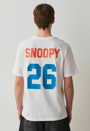 Személy, aki fehér pólót visel, amelyen narancssárgán a "SNOOPY", kék színnel a "26" szerepel a hátoldalon, egyszerű háttér előtt áll.