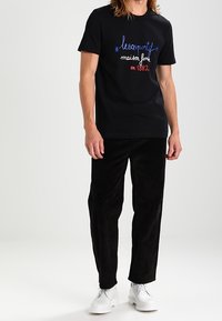 Svart bomull t-shirt med röd och blå broderad text, kombinerad med svarta manchesterbyxor och vita skor. En enkel och modern design.