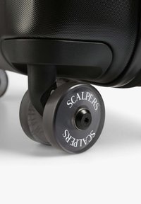 Esquina de equipaje texturizado negro con una rueda transparente marcada "SCALPERS". La rueda presenta un centro negro y una superficie de rodadura lisa.