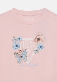 Ροζ βαμβακερό μπλουζάκι με γραφικά λουλουδιών και πεταλούδας, με τα κείμενα "GUESS" και "Los Angeles" σε λευκό και μπλε. Κοντά μανίκια, λαιμόκοψη τύπου crew.