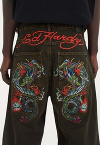 Pantalones de mezclilla verdes y negros con un vibrante bordado de dragón en rojo, azul y blanco en los bolsillos traseros, con "Ed Hardy" bordado en rojo encima.