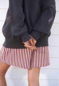 Tmavě šedá oversized mikina s manžetovými rukávy v kombinaci s pruhovanou sukní ke kolenům v červené a krémové barvě. Ruce jsou sepnuté za zády.