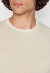 Light beige knit sweater with a round neckline, fine texture, and ribbed cuffs. Close-up view of the upper chest area.