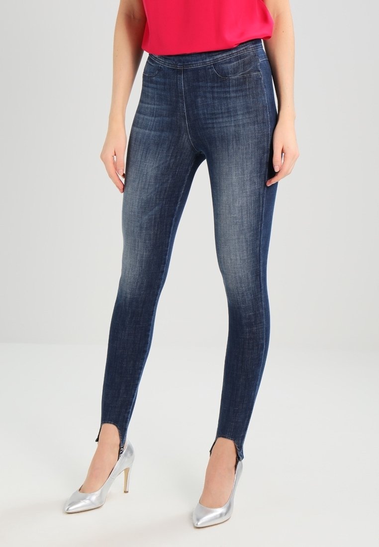 Femme portant un jean skinny bleu foncé avec des poches avant et des fermetures éclair aux chevilles, associé à des talons hauts argentés et un top rouge.
