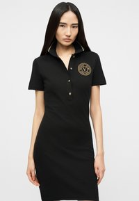 Γυναίκα που φοράει εφαρμοστό μαύρο φόρεμα πόλο με κοντά μανίκια και χρυσό λογότυπο Versace Jeans Couture στο στήθος.