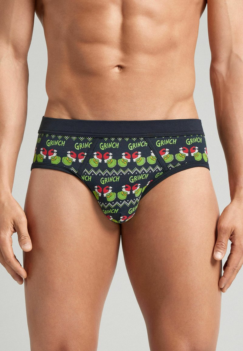 Męski tors w granatowych majkach w wzór z powtarzającymi się zielonymi twarzami Grincha, noszącymi czerwone czapki Mikołaja oraz z napisem "GRINCH" w zielonym kolorze.