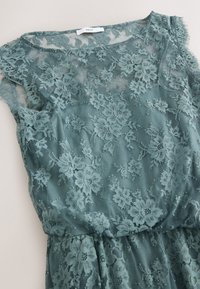 Robe en dentelle bleu sarcelle sans manches avec motifs floraux, corsage supérieur transparent et taille froncée, étendue à plat sur une surface claire.