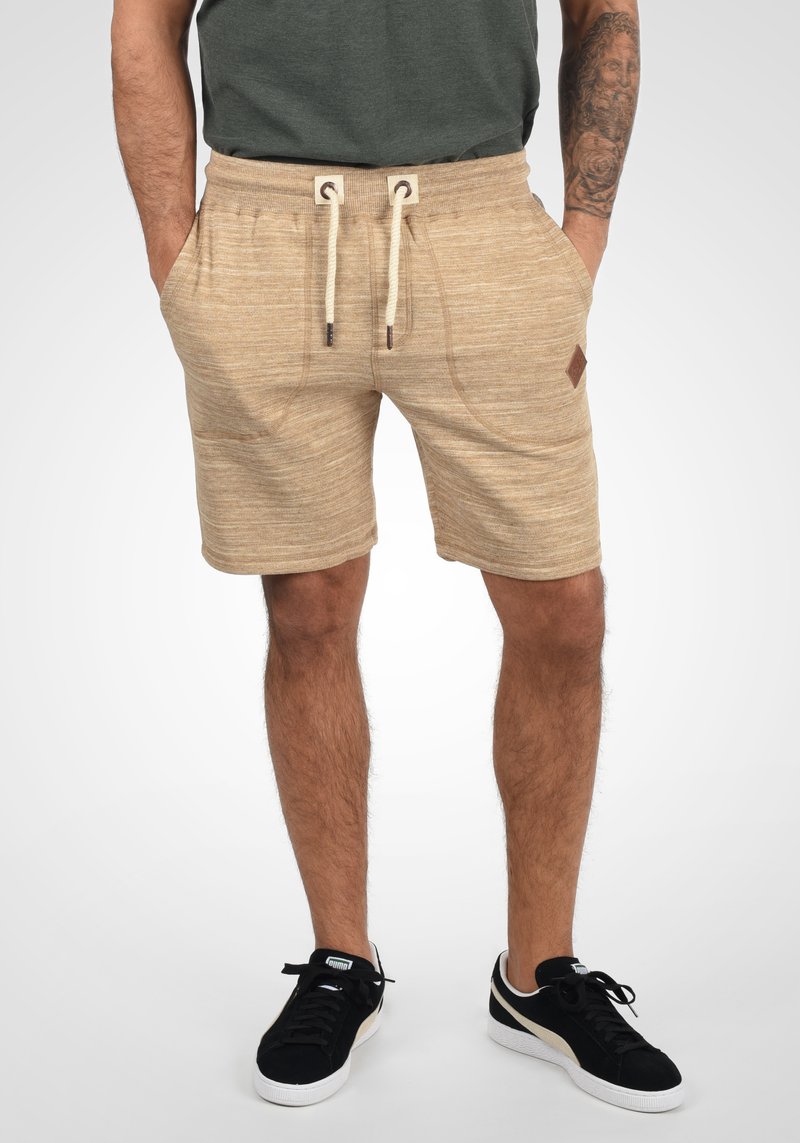 Shorts en tricot beige avec une taille élastique, fermeture à cordon, poches latérales et finition texturée. Accent logo noir sur la taille.