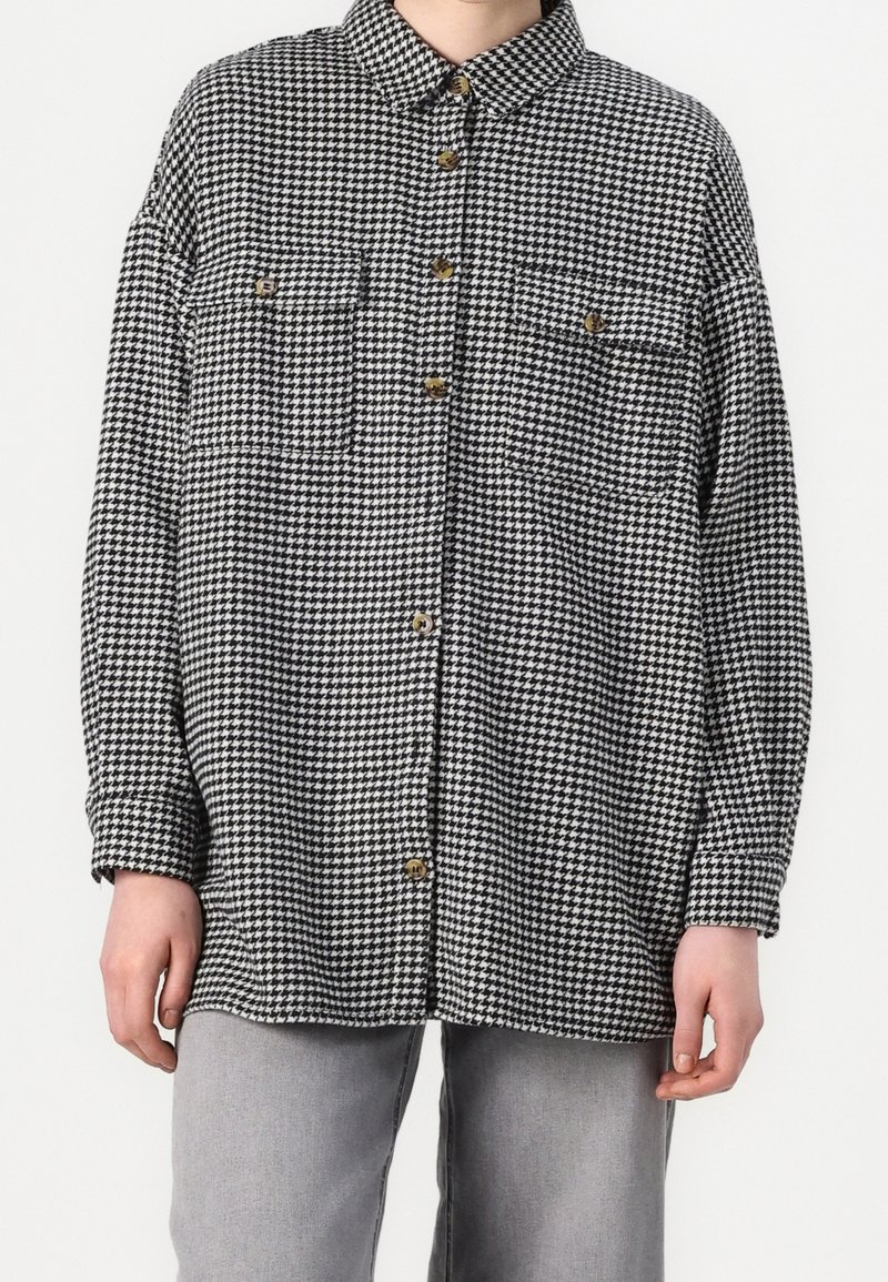 Czarna i biała koszula w wzór houndstooth z kołnierzykiem, dwiema kieszeniami na piersi i zapięciem na guziki, wykonana z teksturowanej tkaniny.