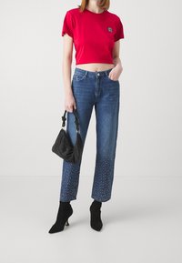 Mujer vestida con un top corto rojo, jeans azules con adornos de perlas en los dobladillos, botas negras de tacón, y sosteniendo un bolso negro con asas anudadas.