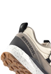 Beige und grauer Sportschuh mit einer strukturierten Oberseite, schwarzen Akzenten und einer gesprenkelten weißen Sohle. Verfügt über ein markantes Logo an der Seite.