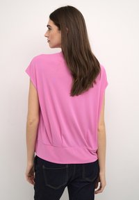 Rosa kortärmad topp med löst passform och samlad bak, tillverkad av mjukt tyg. Matchas med mörka jeans med bakfickor.