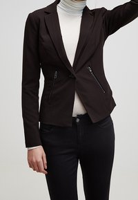 Musta räätälöity bleiseri, jossa on läpät, kaksi vetoketjullista etutaskua ja muotoillut saumat. Pukeutuneena vaalean turtleneckin päälle.