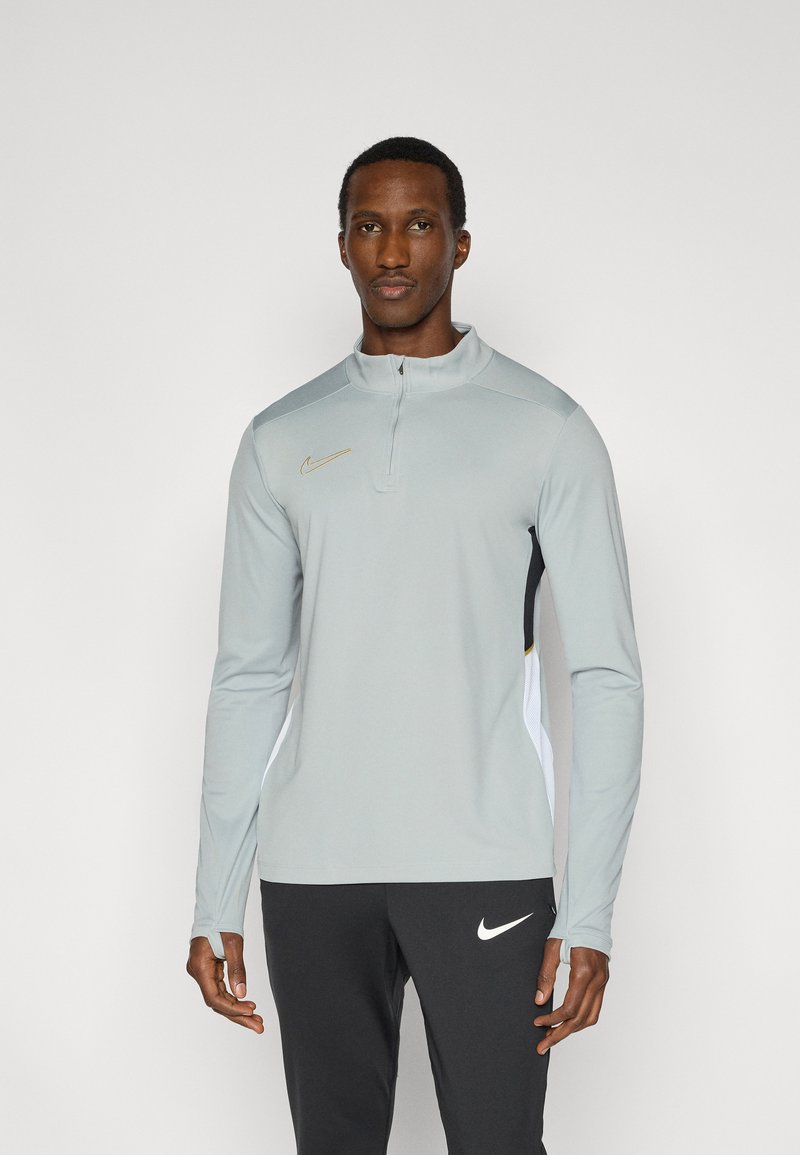 Ανοιχτό γκρι μπλούζα με μακριά μανίκια της Nike, με λαιμόκοψη μισό φερμουάρ, που διαθέτει μαύρες και λευκές πλευρικές πλάκες και χρυσές λεπτομέρειες. Λείο ύφασμα.