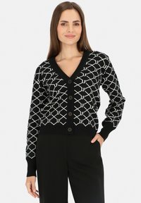 Czarny kardigan z geometrycznym białym wzorem, dekoltem w serek oraz pięcioma ciemnymi guzikami z przodu. Żebrowane mankiety i dół dodają tekstury.