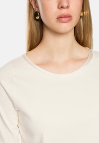 Cream-colored short-sleeve top with a round neckline, smooth texture, and visible stitching detail. Model wears large gold circular earrings.