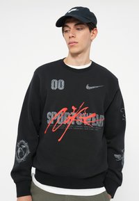 Črna majica s kapuco z večbarvnimi grafičnimi elementi, ki vključujejo rdeče in sivo besedilo. Vsebuje Nike logo, imena različnih mest in podrobne poudarke na rokavih.
