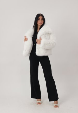 Frau mit langen dunklen Haaren, die eine weiße Kunstpelzjacke über einem schwarzen Outfit und weiße offene Absätze trägt, steht vor einem schlichten Hintergrund.