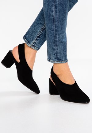 Black suede heeled shoes with a pointed toe and open back, featuring a chunky heel and curved upper design, paired with faded blue jeans.