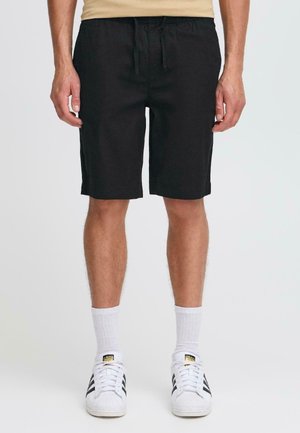 Homme portant un short noir jusqu’aux genoux, des chaussettes blanches et des baskets blanches Adidas avec des rayures noires, debout devant un fond uni.