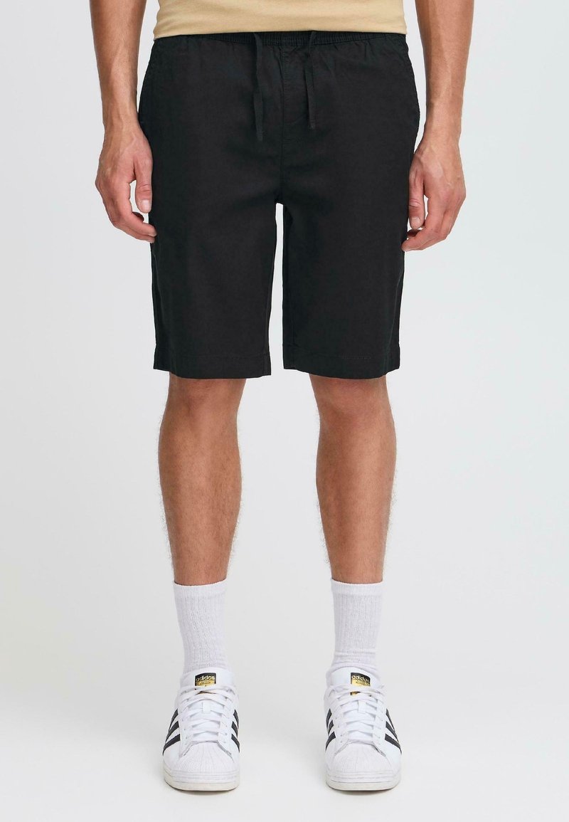 Homme portant un short noir jusqu’aux genoux, des chaussettes blanches et des baskets blanches Adidas avec des rayures noires, debout devant un fond uni.