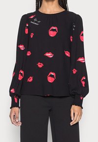 Svart långärmad blus med mönster av röda läppar och vit text "life is awesome", kombinerad med svarta byxor, händer avspända vid sidorna.