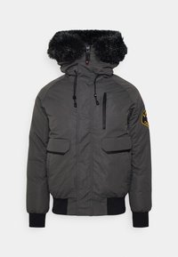 Grå polstret jakke med hætte foret med kunstpels, elastiske manchetter, to frontlommer og en lynlåslomme på brystet. Sorte detaljer er til stede.