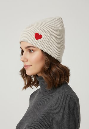 Cream ribbed knit beanie with a red heart patch. Features a tapered top and a folded brim. Worn with a dark gray ribbed turtleneck.