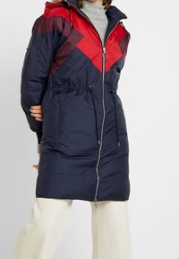 Abrigo acolchado de la marina con patrón en forma de chevron rojo, corte regular, cierre de cremallera, cinturilla ajustable con cordón y capucha. Textura suave y lisa.