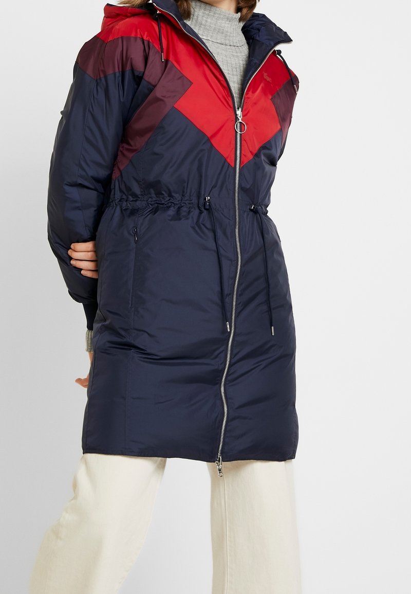 Abrigo acolchado de la marina con patrón en forma de chevron rojo, corte regular, cierre de cremallera, cinturilla ajustable con cordón y capucha. Textura suave y lisa.