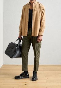 Beige Langarmhemd, schwarzes Tanktop, olivgrüne Hose, schwarze Schuhe und eine schwarze Sporttasche. Holzboden und hellgrauer Hintergrund.