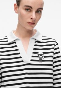 Schwarz-weißer, gestreifter Poloshirt mit V-Ausschnitt, der ein kleines illustriertes Charakterdesign auf der linken Seite zeigt. Das Material scheint reich an Baumwolle zu sein.