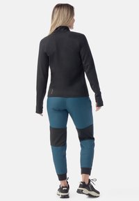 Giacca in pile nera con zip abbinata a pantaloni da jogging teal e neri, caratterizzati da un design a pannelli. Indossata con sneakers nere.
