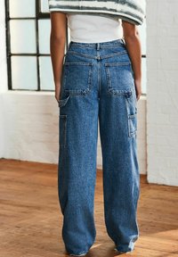 Jeans din denim albastru cu talie înaltă și picioare late, având buzunare mari pe spate și pe laterale, cu un tiv brut și o textură estompată.