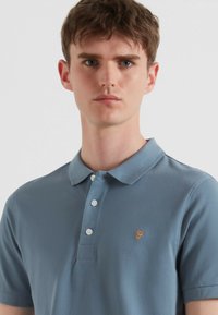 Lichtblauw polo-shirt gemaakt van textuurstof, met een klassieke kraag, drie witte knopen en een oranje embleem op de linker borst.