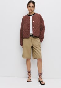 Jachetă bomber căptușită maroon, tricou alb, pantaloni scurți bej, curea neagră, sandale negre cu șireturi multicolore. Ținută simplă și casual.