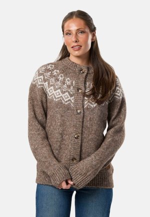 Vrouw met lang bruin haar, draag een bruine gebreide cardigan met witte geometrische patronen over de schouders, gecombineerd met een blauwe spijkerbroek.