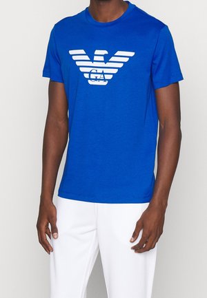 Hombre con una camiseta de manga corta de color azul brillante con un gran logo blanco de águila y pantalones blancos sobre un fondo liso.