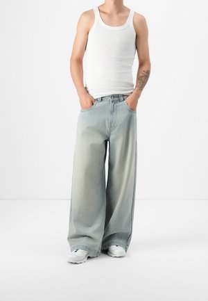 Personne portant un débardeur blanc, un jean large et ample bleu clair, des baskets blanches, les mains dans les poches et un tatouage sur l'avant-bras gauche.