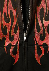 Sweat à capuche noir zippé arborant de grands motifs de flammes rouges avec des accents jaunes. Le tissu semble doux, avec un détail de fermeture éclair argentée brillante.