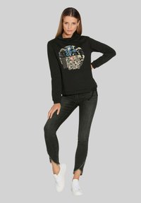 Schwarzes langärmeliges Sweatshirt mit Grafikdruck, das verschiedene Muster zeigt. Kombiniert mit dunkelgrauen Jeans und weißen Sneakern.