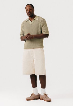 Mann som står med sammenflettede hender, iført beige strikket kortermet genser, lyse shorts, hvite sokker og beige snøresko på hvit bakgrunn.